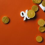 Gold coins scattered with a stock market graph and a percentage symbol on an orange background.
