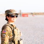 Adult female soldier in camouflage uniform smiling confidently while standing outdoors on a bright day.