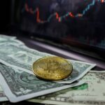 A bitcoin coin placed on US dollar bills with a stock market chart in the background.