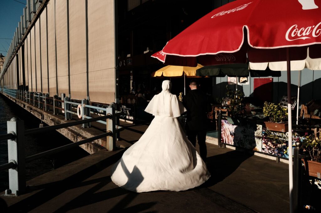 A bride and groom walk along the Istanbul waterfront, casting long shadows in the evening sun.