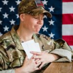 A soldier in uniform holds envelopes emotionally in front of an American flag background.