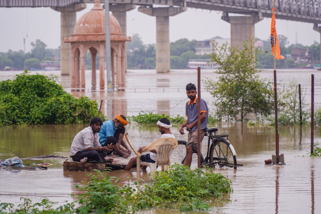 A group of people gather by the flooded river in Varanasi, with a bridge and temple visible.