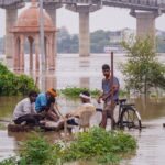 A group of people gather by the flooded river in Varanasi, with a bridge and temple visible.
