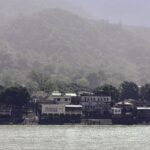 View of a tranquil riverside village with dense green hills in the background, ideal for travel and nature scenes.