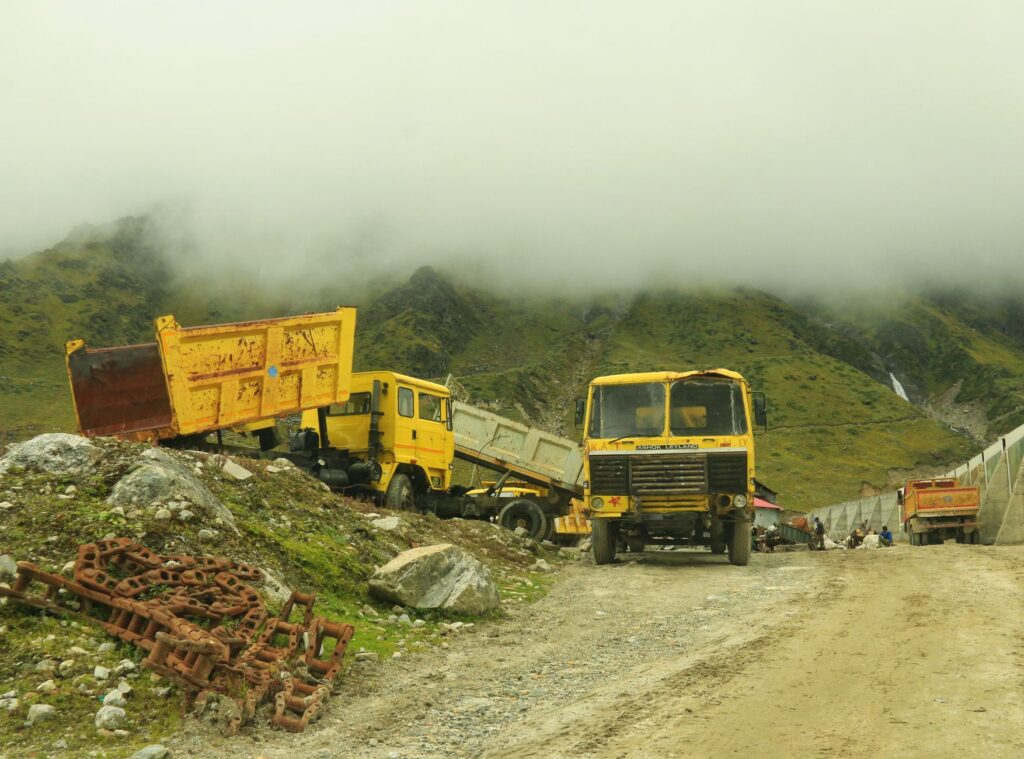 Yellow industrial trucks on a misty road in Kedarnath, India, amid rocky terrain and mountains.