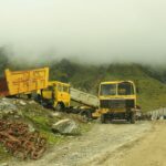 Yellow industrial trucks on a misty road in Kedarnath, India, amid rocky terrain and mountains.