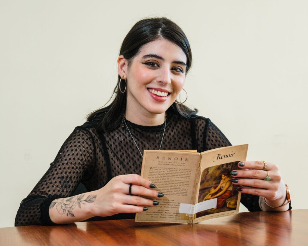 Young woman with tattoos reading a Renoir book indoors, smiling.