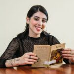 Young woman with tattoos reading a Renoir book indoors, smiling.