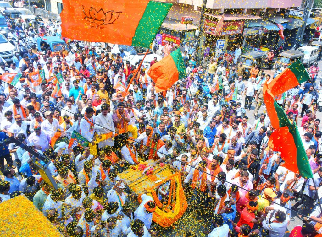 Crowds gather in Mangaluru for a colorful street parade with vibrant flags and decorations.