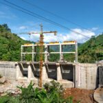 Crane operating at a construction site in a lush, mountainous area under bright blue skies.