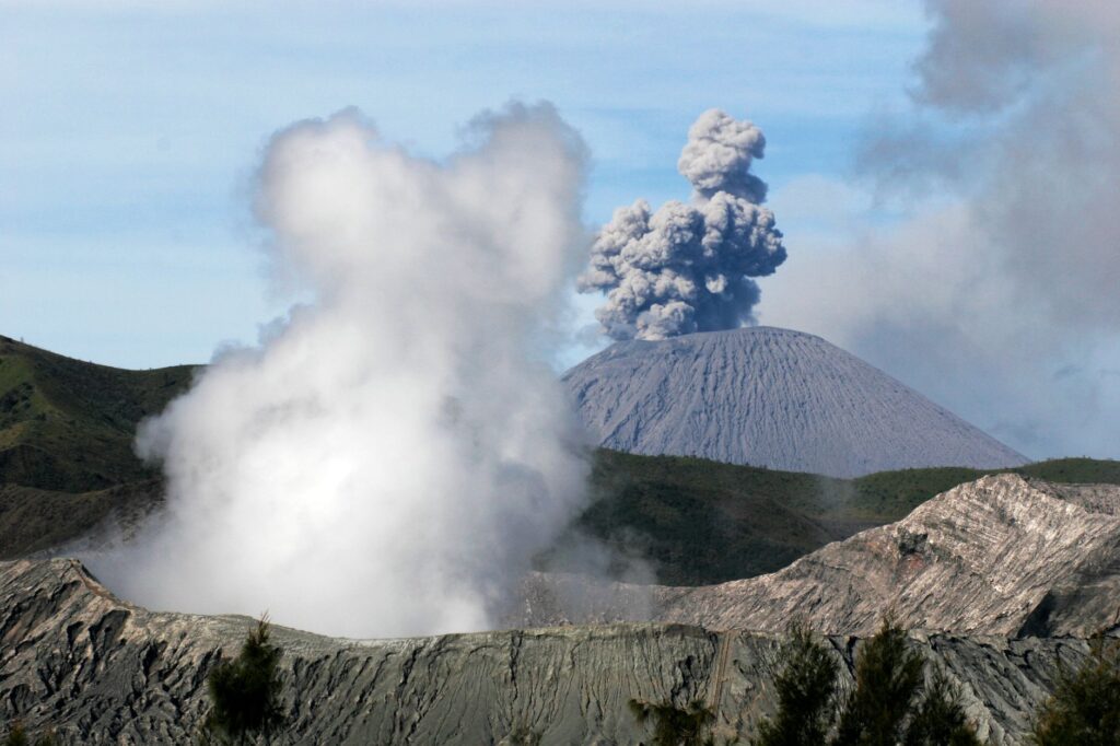 A dramatic aerial view of an erupting volcano with thick smoke plumes and rugged landscape.