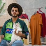 Man in hat with a cat and hanging shirts in a relaxed outdoor setting.