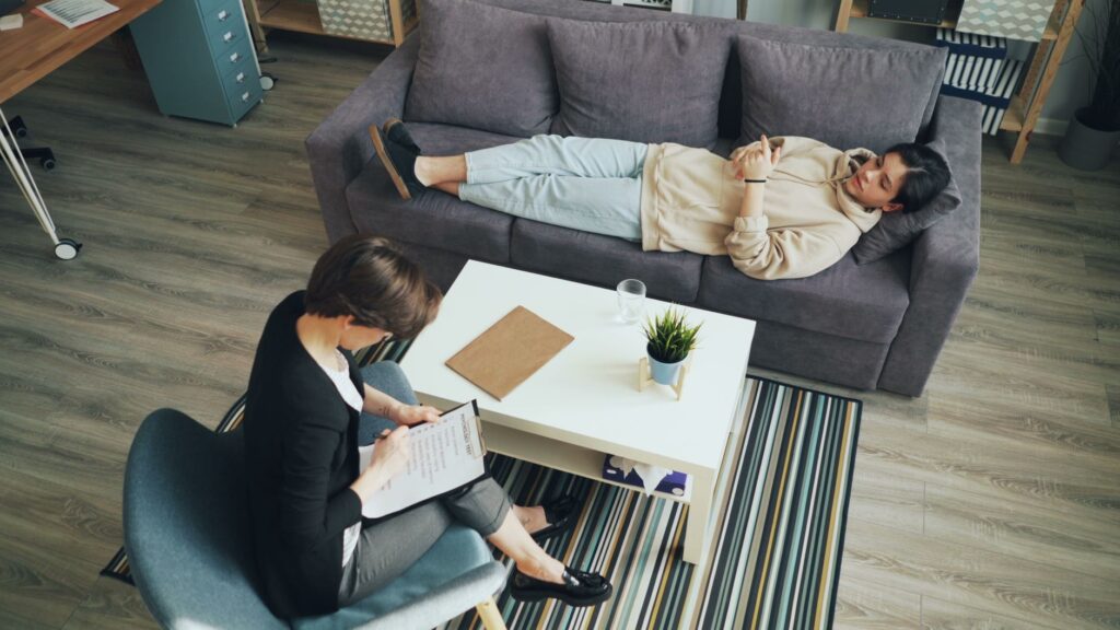 A therapist consults with a client lying on a couch in a modern office setting.