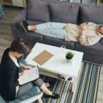 A therapist consults with a client lying on a couch in a modern office setting.