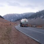 Gas tanker truck traveling on a mountainous road with snow-capped hills in the background.