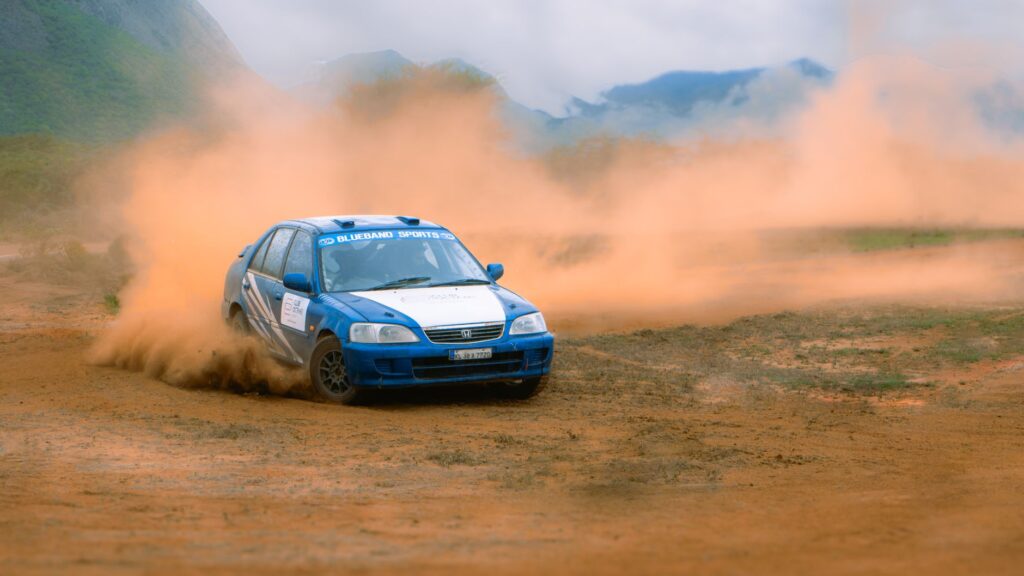 Blue rally car racing on a dirt track with dust clouds.