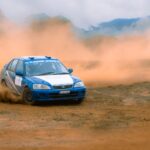 Blue rally car racing on a dirt track with dust clouds.