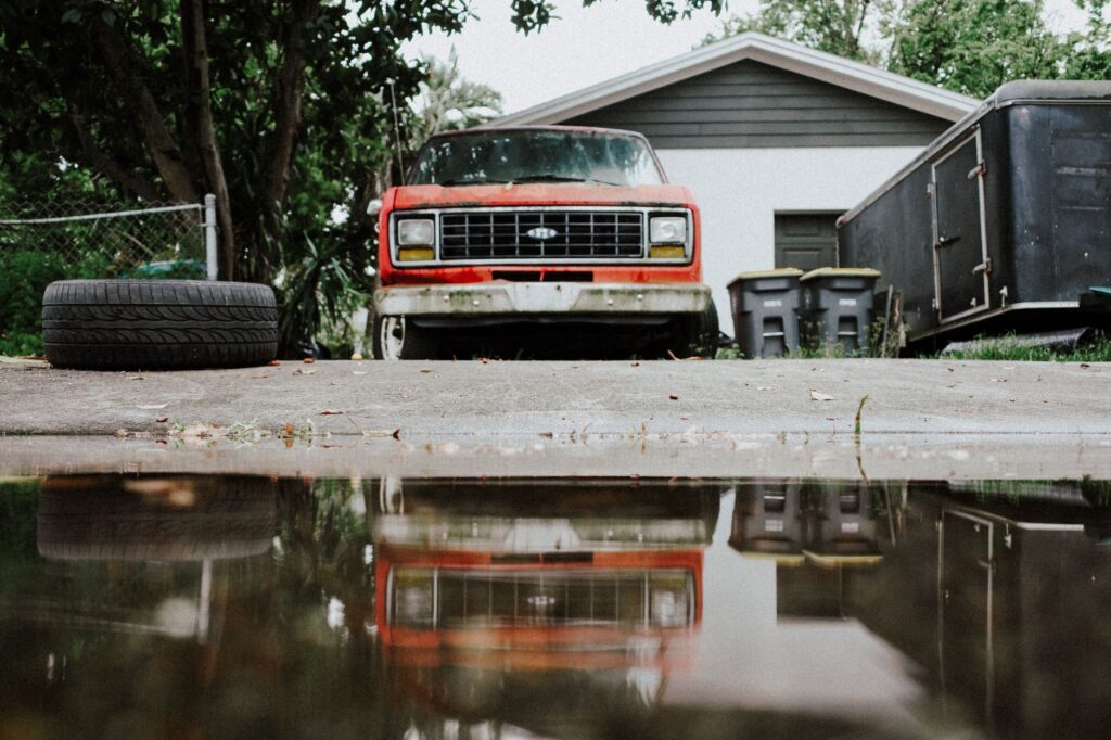 Old red truck with reflection on a wet driveway, showcasing rustic charm.