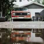 Old red truck with reflection on a wet driveway, showcasing rustic charm.