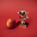 A 'Best Teacher' trophy alongside an apple on a vivid red background symbolizing education and recognition.