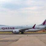 Qatar Airways aircraft captured on runway at Tbilisi airport. Clear sky and distant hills enhance the scene.
