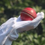 Detailed photo of a red cricket ball held by gloved hands outdoors.