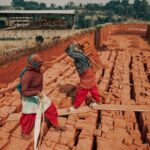 Two workers carrying bricks at an outdoor construction site, showcasing teamwork and labor.