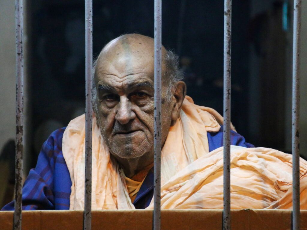 Portrait of an elderly man behind bars wearing casual clothing, conveying a thoughtful mood.