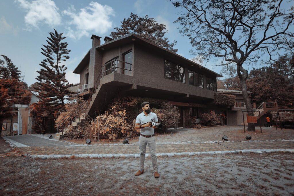 Man standing with camera in front of modern house surrounded by autumn trees and garden.