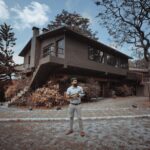 Man standing with camera in front of modern house surrounded by autumn trees and garden.