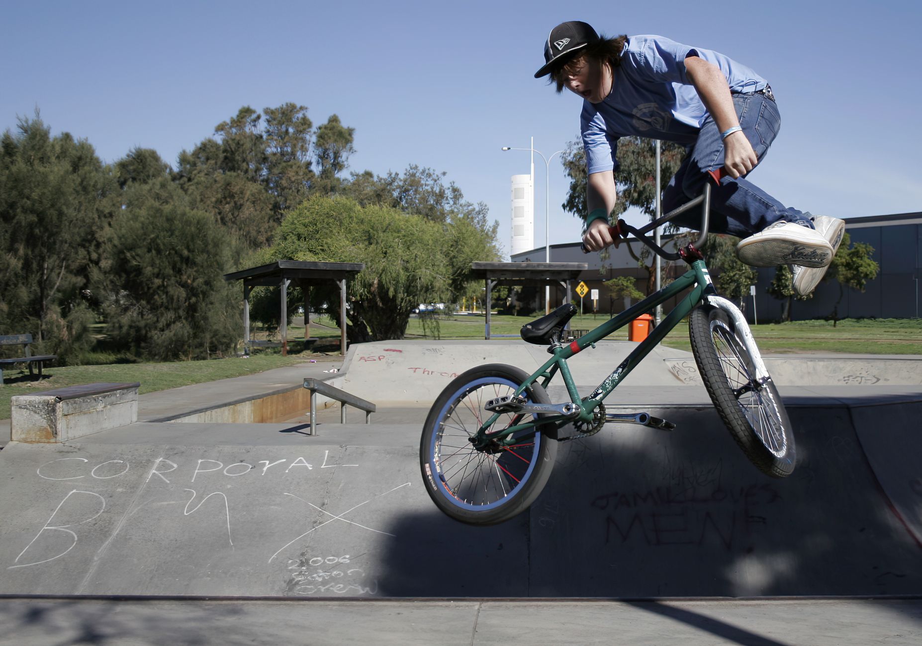 BMX rider performing mid-air trick at a sunny skatepark with green trees and graffiti.