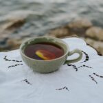 A ceramic cup of tea with a lemon slice, elegantly placed on a lace cloth by a serene lakeside.