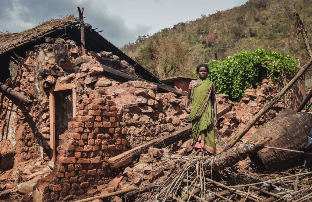 Woman stands in front of a destroyed house after a disaster in rural India, depicting struggle.