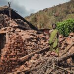 Woman stands in front of a destroyed house after a disaster in rural India, depicting struggle.