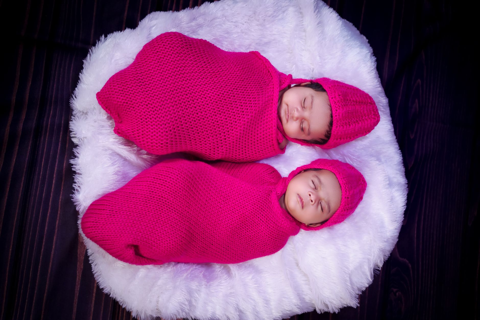Charming image of twin babies peacefully sleeping in pink knit swaddles on a fluffy surface.