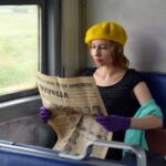 Concentrated young woman wearing elegant gloves and black blouse sitting in train with hand on suitcase and reading old Russian newspaper