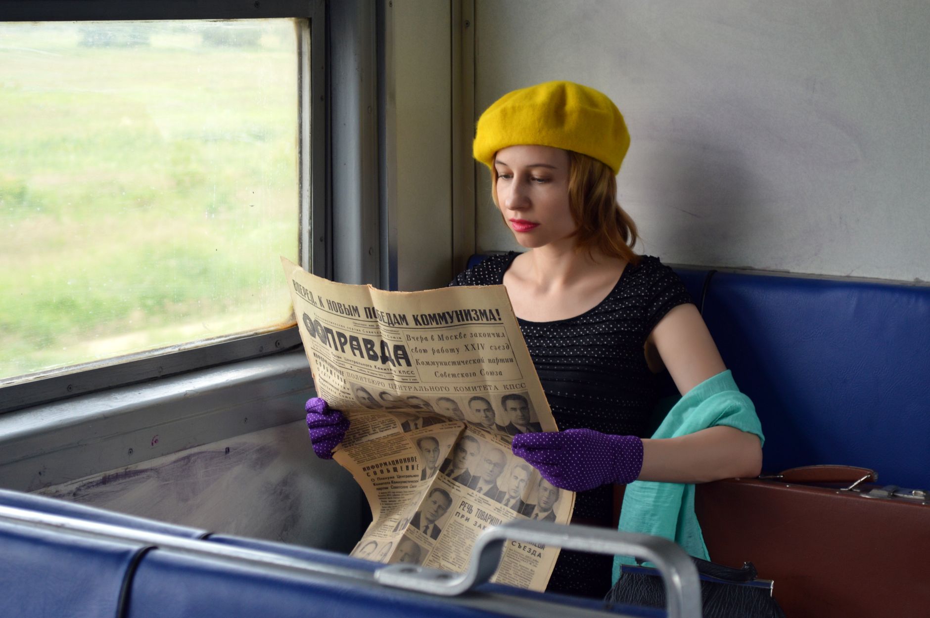 Concentrated young woman wearing elegant gloves and black blouse sitting in train with hand on suitcase and reading old Russian newspaper