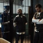 A police officer stands by two prisoners in jail uniforms behind bars, indoors.