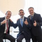 Three businessmen in suits smiling and giving thumbs up, symbolizing team success and positivity.