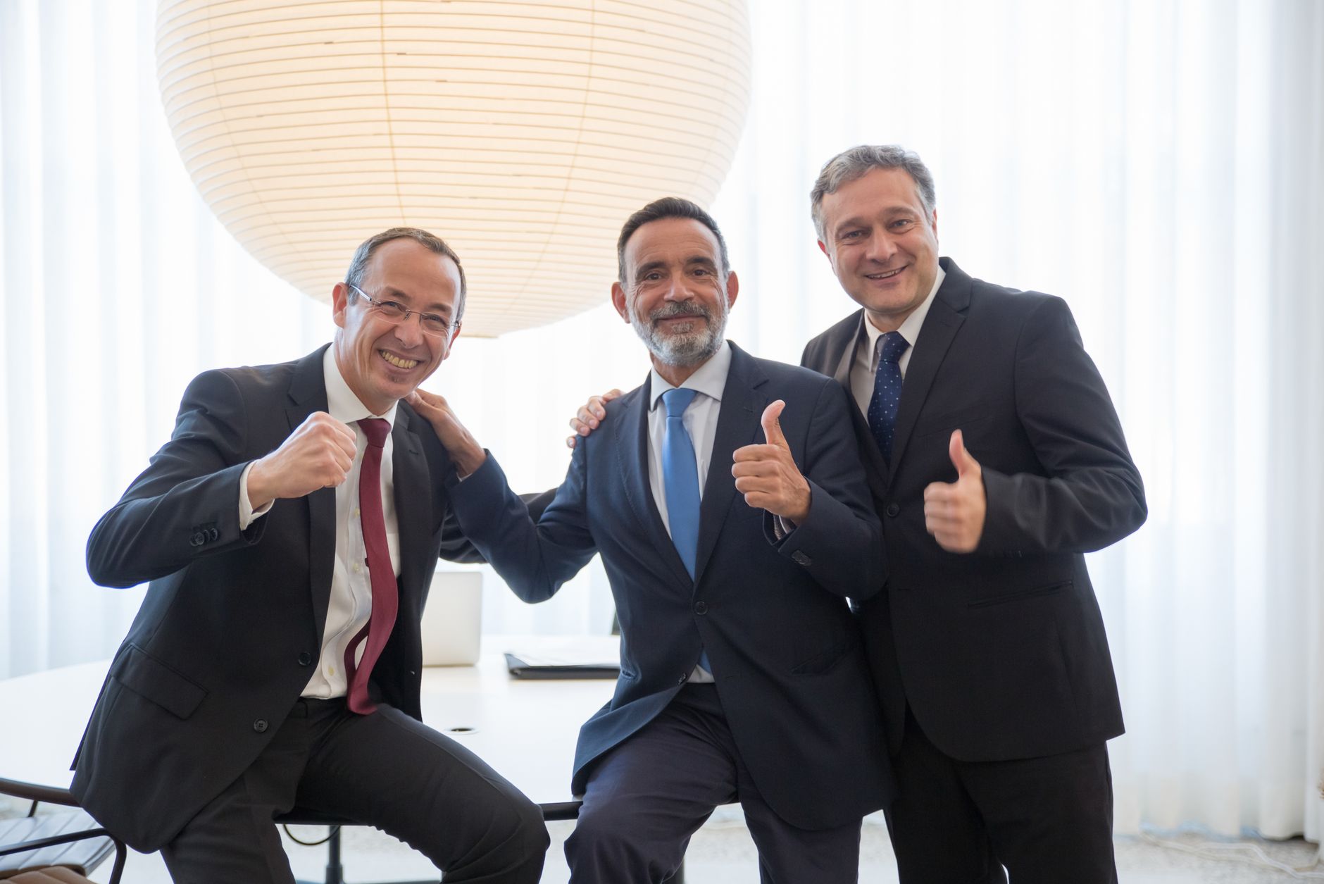 Three businessmen in suits smiling and giving thumbs up, symbolizing team success and positivity.