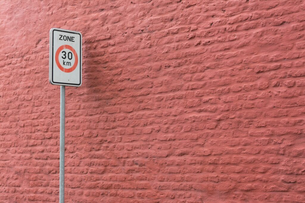 A 30 km/h speed limit sign against a textured pink brick wall in an urban setting.
