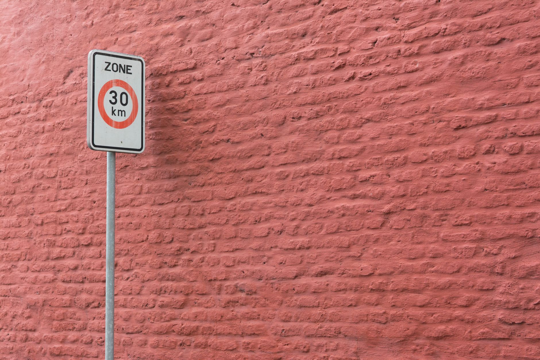 A 30 km/h speed limit sign against a textured pink brick wall in an urban setting.
