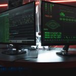 Close-up of dual computer monitors with green coding interfaces in a dark room, highlighting cyber security themes.