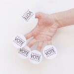 A hand displays several 'Vote' stickers on a white background, symbolizing political participation.