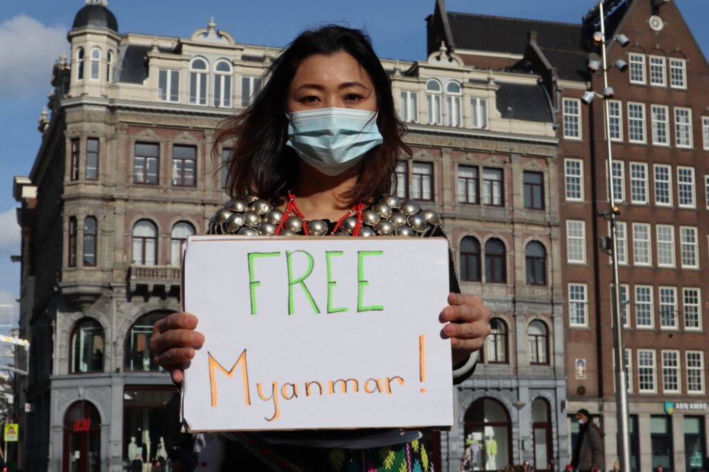 Woman protesting with a sign for Myanmar freedom in urban backdrop, wearing a mask.
