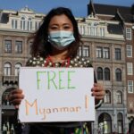 Woman protesting with a sign for Myanmar freedom in urban backdrop, wearing a mask.