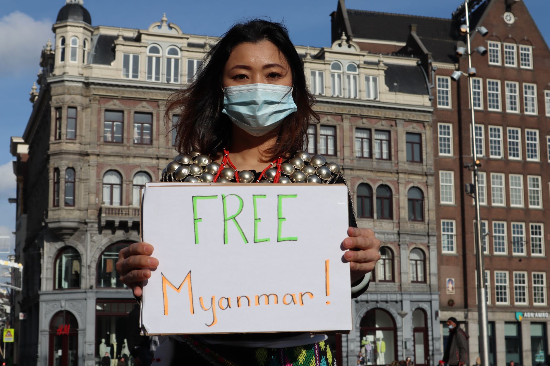 Woman protesting with a sign for Myanmar freedom in urban backdrop, wearing a mask.