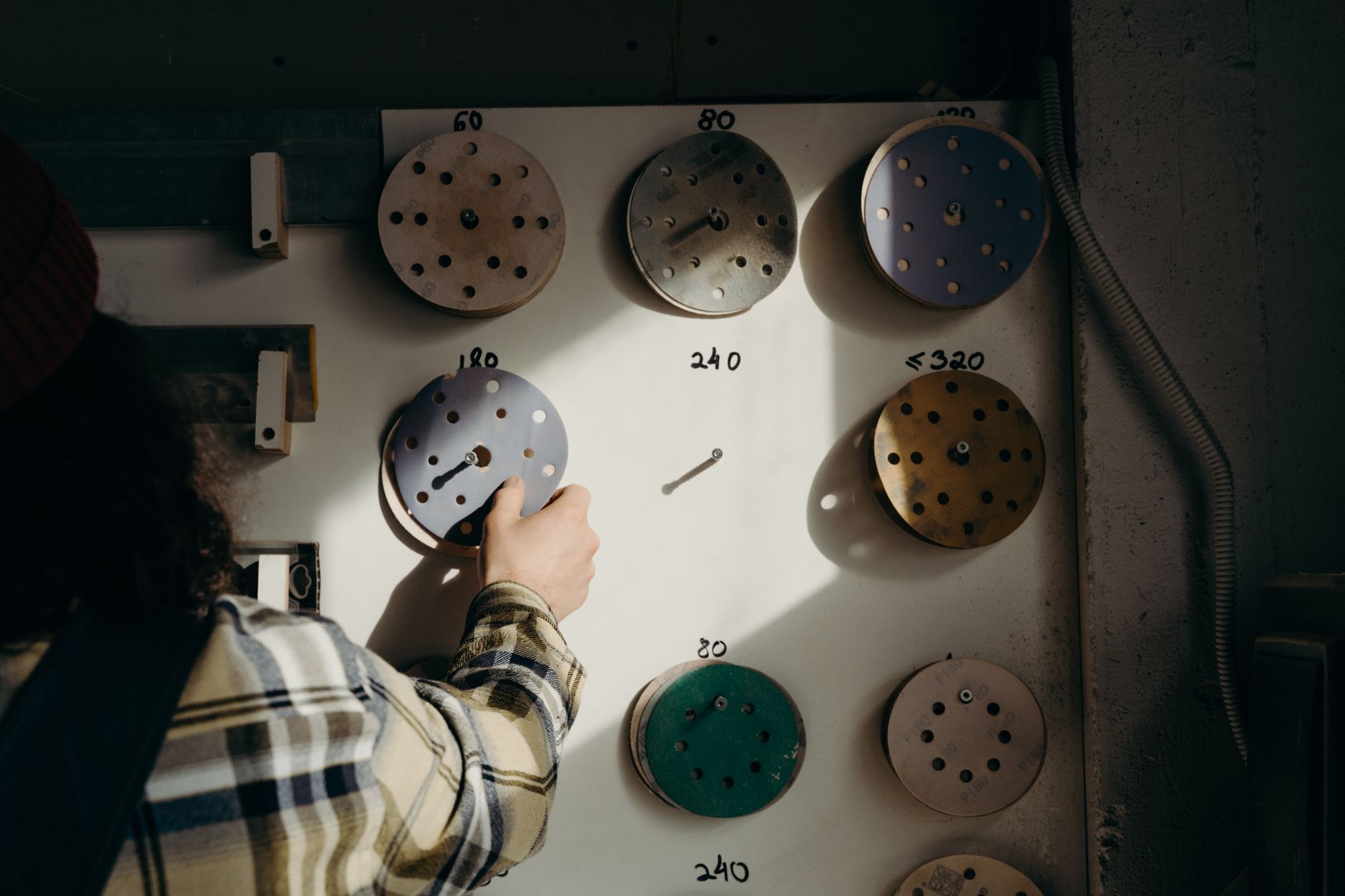 A person in a carpentry workshop selects sandpaper disks from a board in warm lighting.