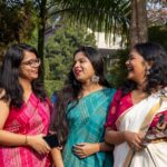 A joyful trio of women in traditional Indian sarees sharing a candid moment outdoors.