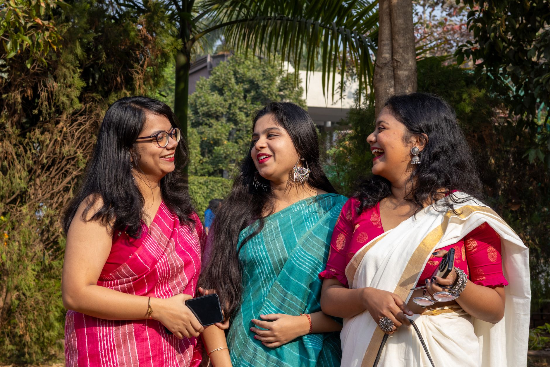 A joyful trio of women in traditional Indian sarees sharing a candid moment outdoors.